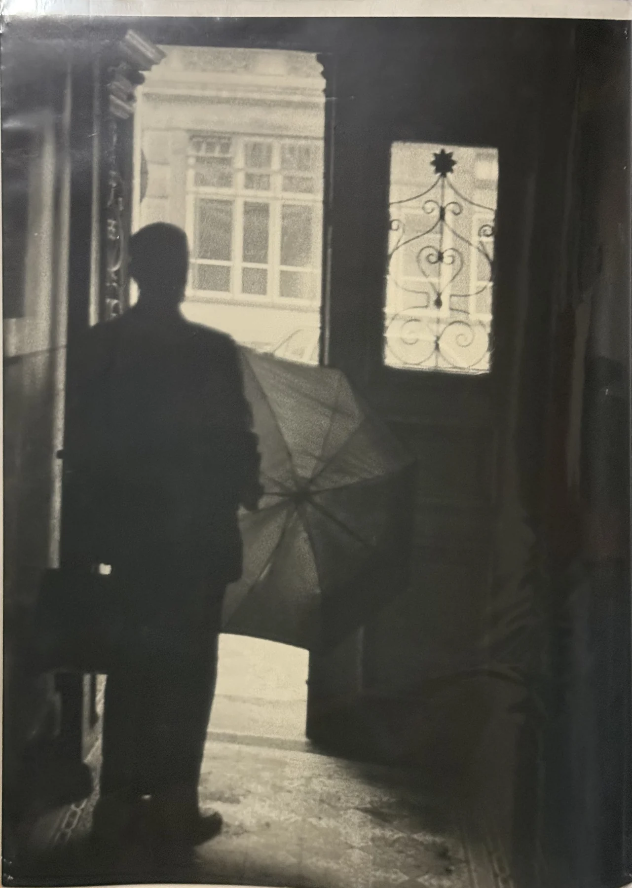 Eine Silhouette steht am Übergang zu einer hellen Öffnung. Gemustertes Glas und ein Schirm gliedern den dunklen Innenraum.