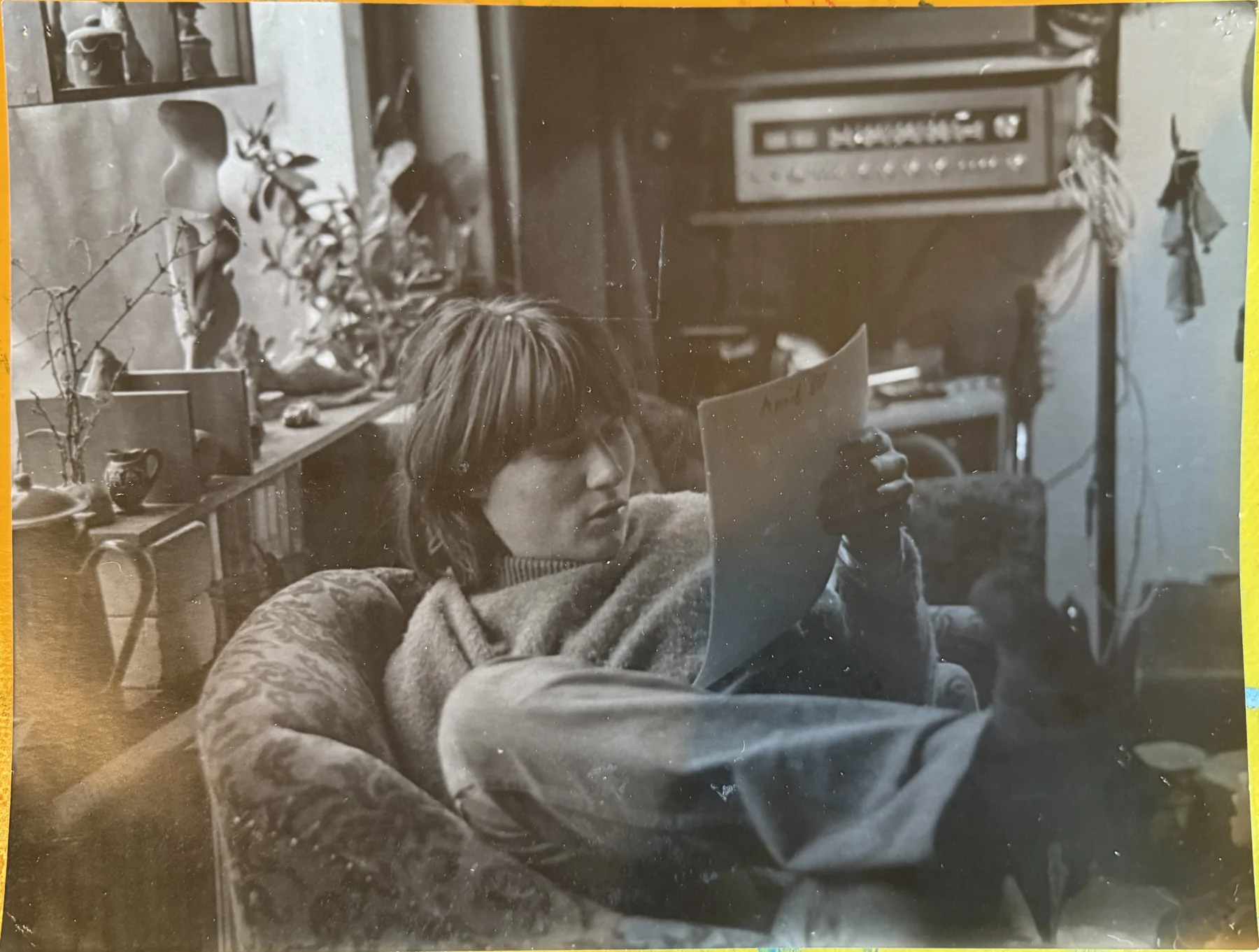 Michael Plötz sitzt mit einem aufgeschlagenen Buch oder einer Zeitschrift zwischen Pflanzen, gemustertem Stoff und Audiogeräten.